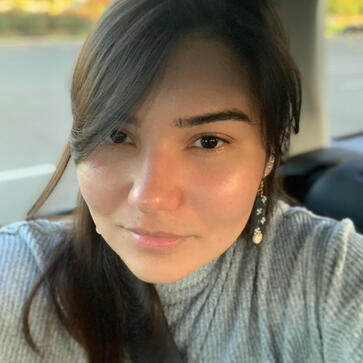 Photo of Kay. She has dark brown hair, flowing to one side and has a slight smile.